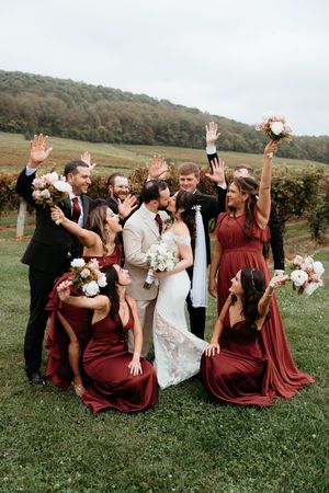 Wedding party in burgundy dresses poses playfully on grassy hillside with white bouquets.