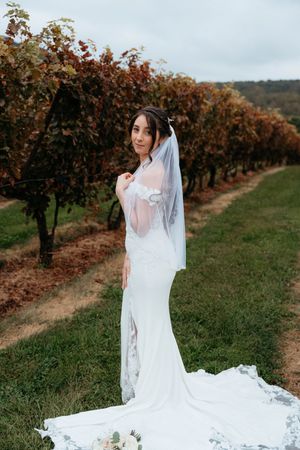 A bride in a white lace gown stands elegantly in a vineyard with autumn foliage in the background.