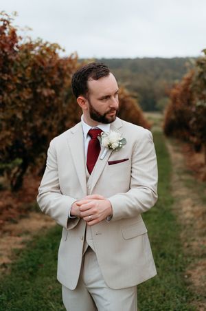A groom in a light beige suit with burgundy tie stands thoughtfully in an autumn vineyard setting.