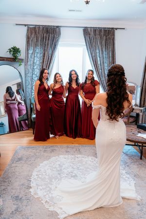 A group in burgundy bridesmaid dresses stands with a bride in a white gown with a flowing train during wedding preparations.