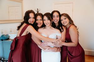 A group of bridesmaids in burgundy dresses embrace and laugh together during a wedding celebration.