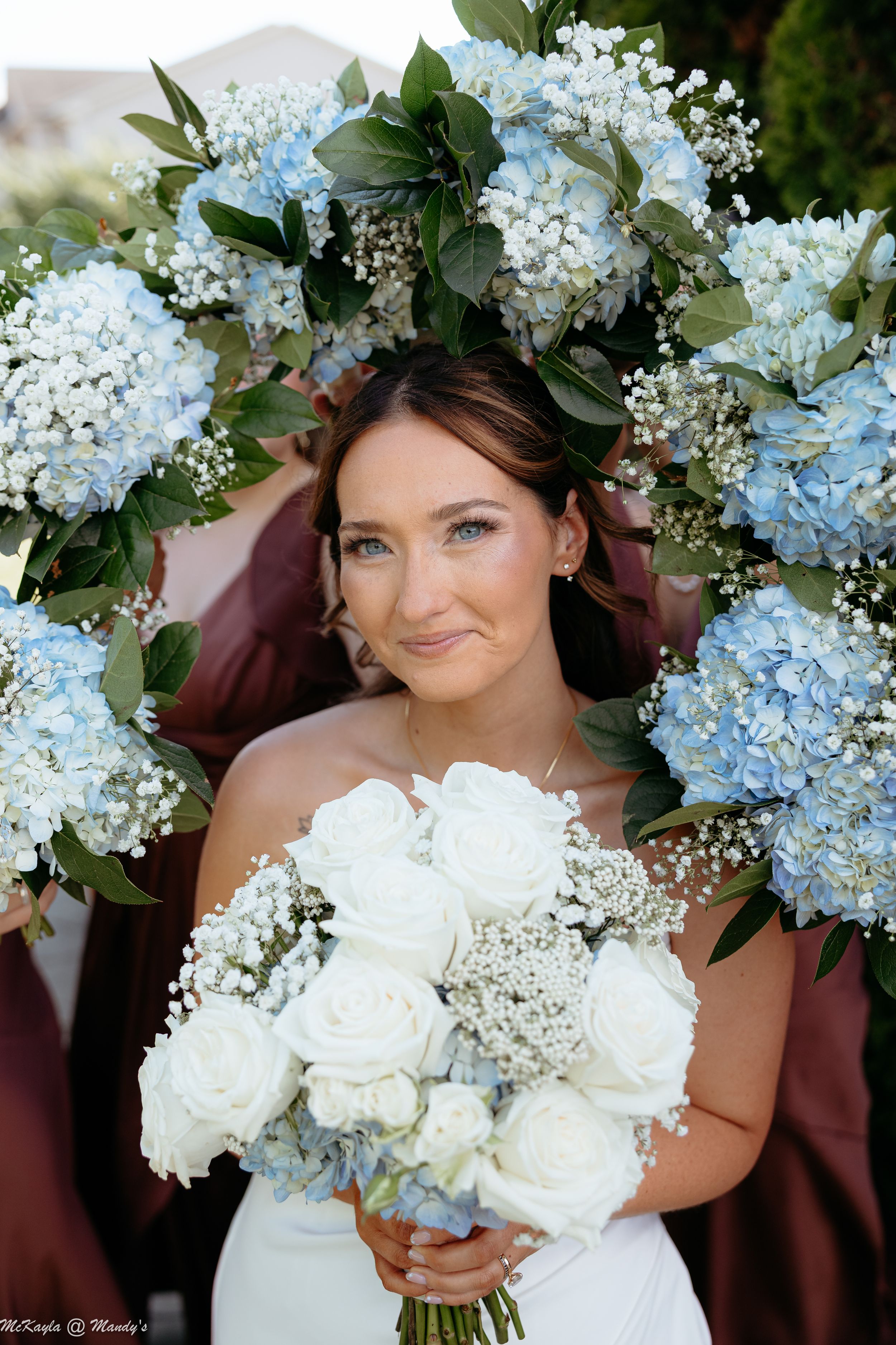Bride holding white rose bouquet surrounded by bridesmaids with blue hydrangea and baby's breath bouquets at wedding.