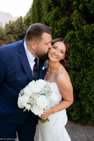 Groom in navy suit kisses bride on cheek as she smiles holding white rose bouquet outdoors.