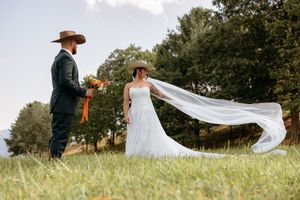Bride in flowing white gown and veil blowing in wind stands with groom in cowboy hat holding bouquet in green field.
