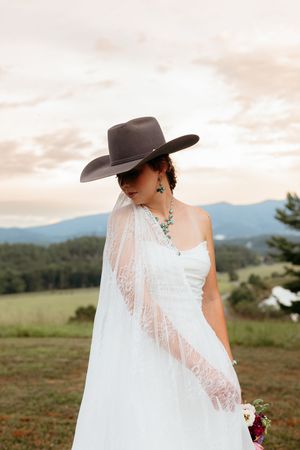 Bride in white lace wedding dress and gray cowboy hat with veil, standing in scenic mountain field at sunset.