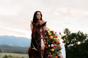 A woman in a white dress rides a brown horse adorned with colorful flowers, set against a mountain backdrop.