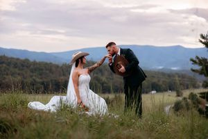 Bride in white gown and cowboy hat sits in meadow as groom in suit serenades her with guitar, mountains in background.