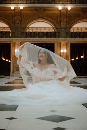 Bride in off-shoulder lace gown with flowing veil in elegant gold-lit grand hall with marble floors.
