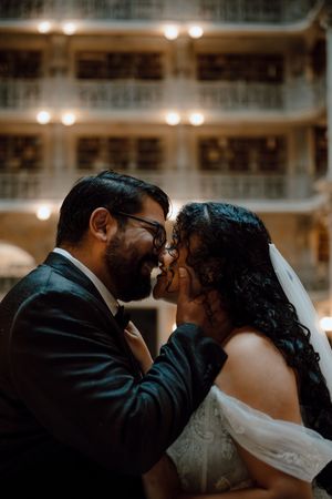 A bride and groom share an intimate moment, nearly kissing, in an elegant warmly lit venue.