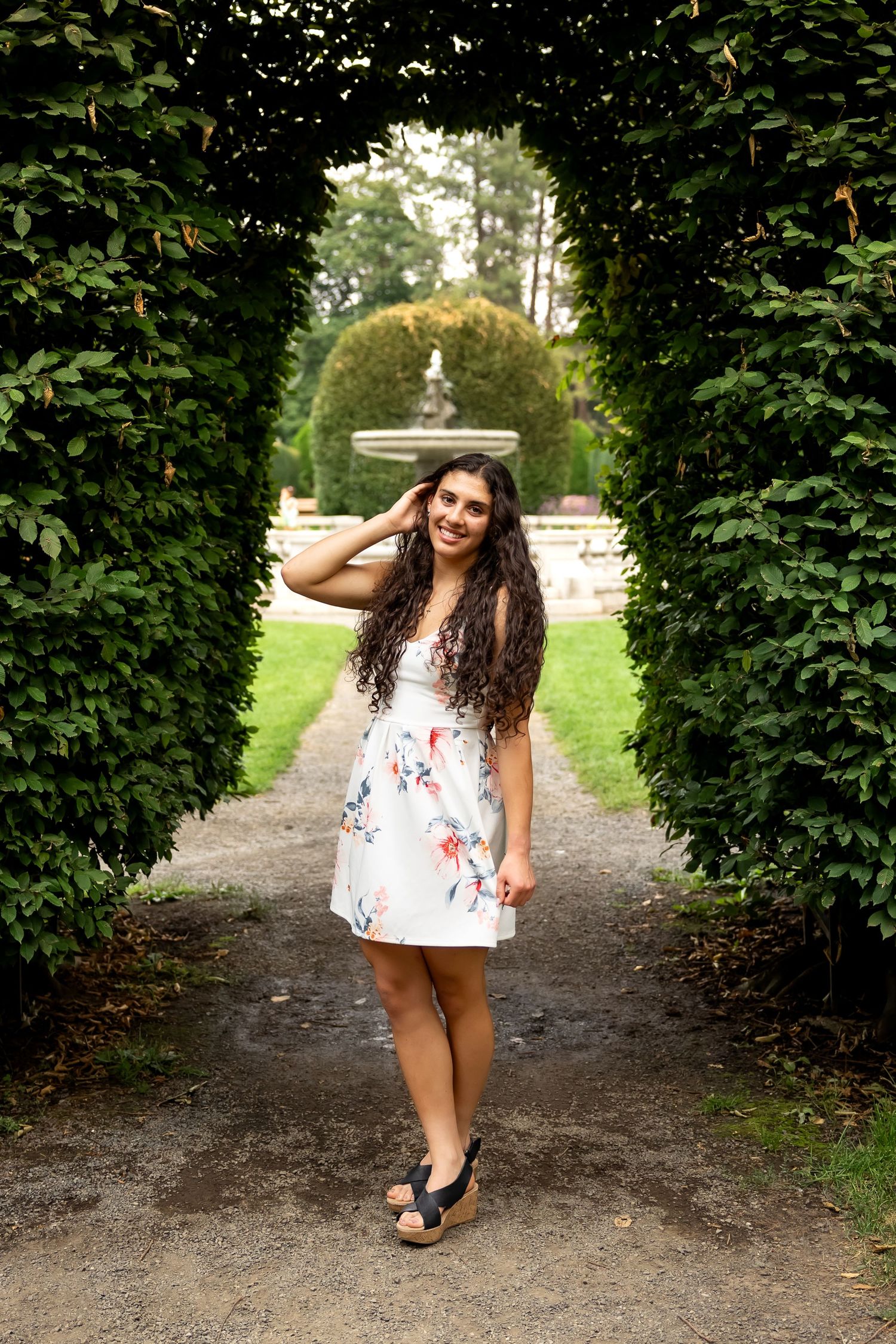 A summer portrait on a garden path with an arched hedge tunnel and fountain in the background.