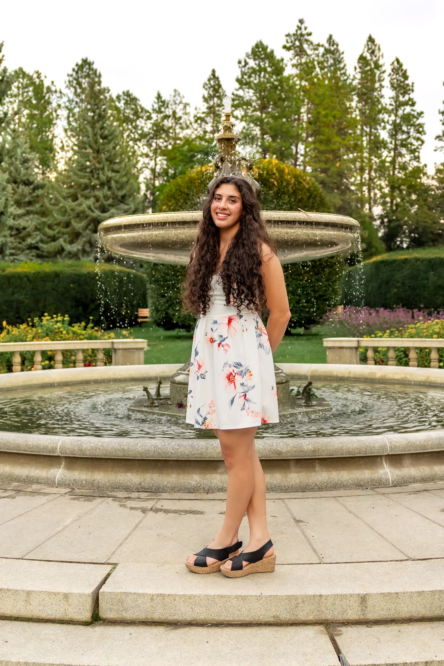 A person in a floral white dress poses near a decorative stone fountain in a garden setting.