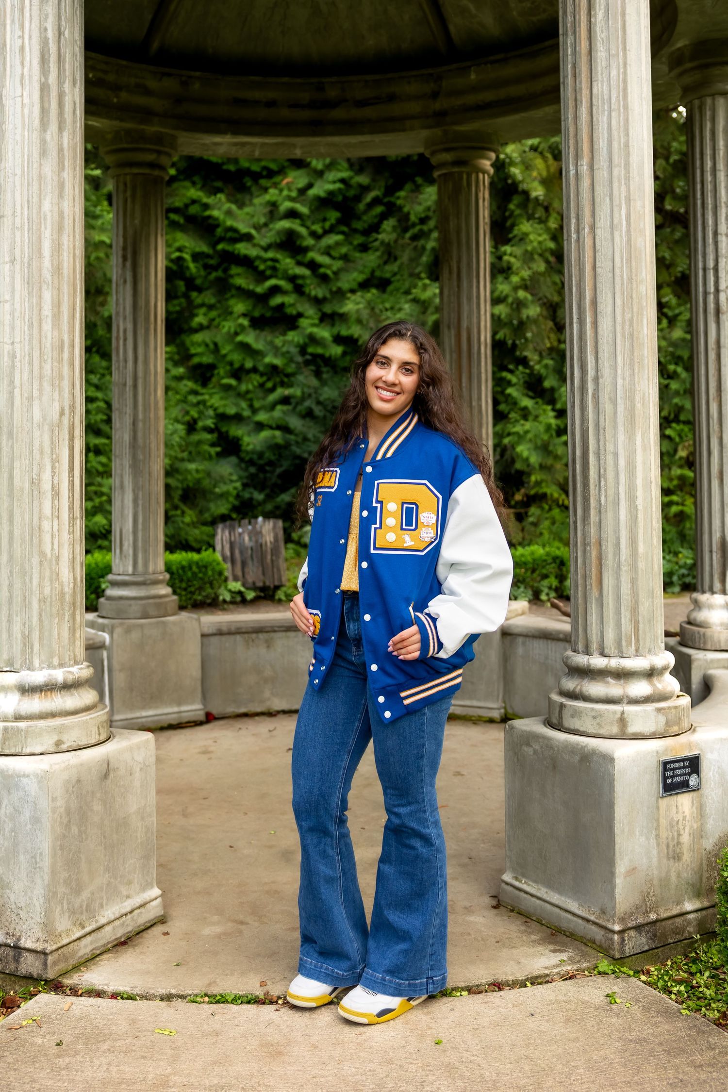 Varsity letterman jacket and flared jeans outfit photographed in classical stone gazebo with green foliage backdrop.