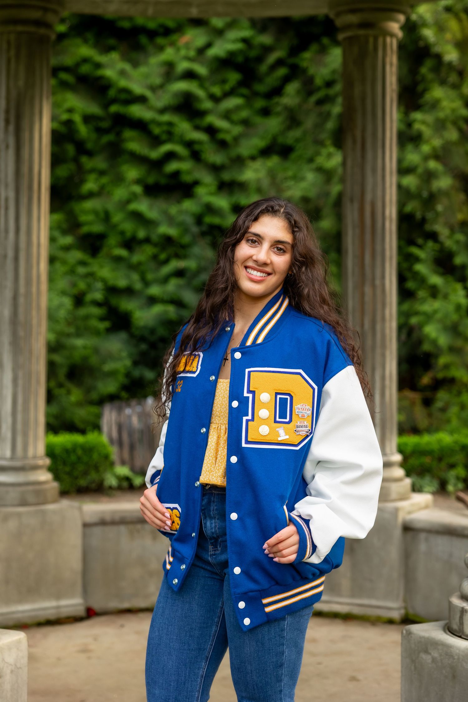 Someone wearing a blue and white varsity letterman jacket poses near stone columns in a garden setting.