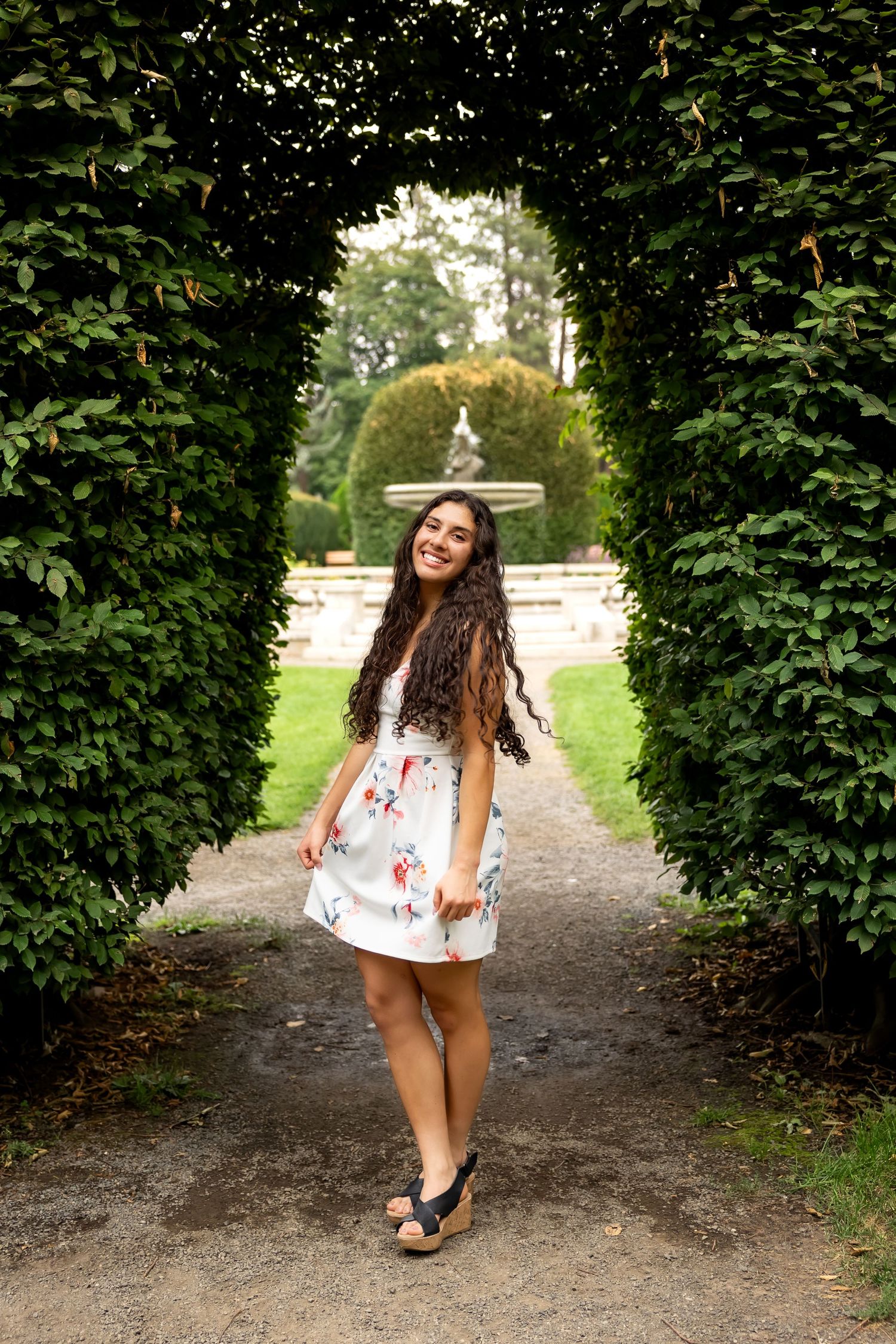 A person in a white top poses playfully on a garden path lined with lush green hedges.