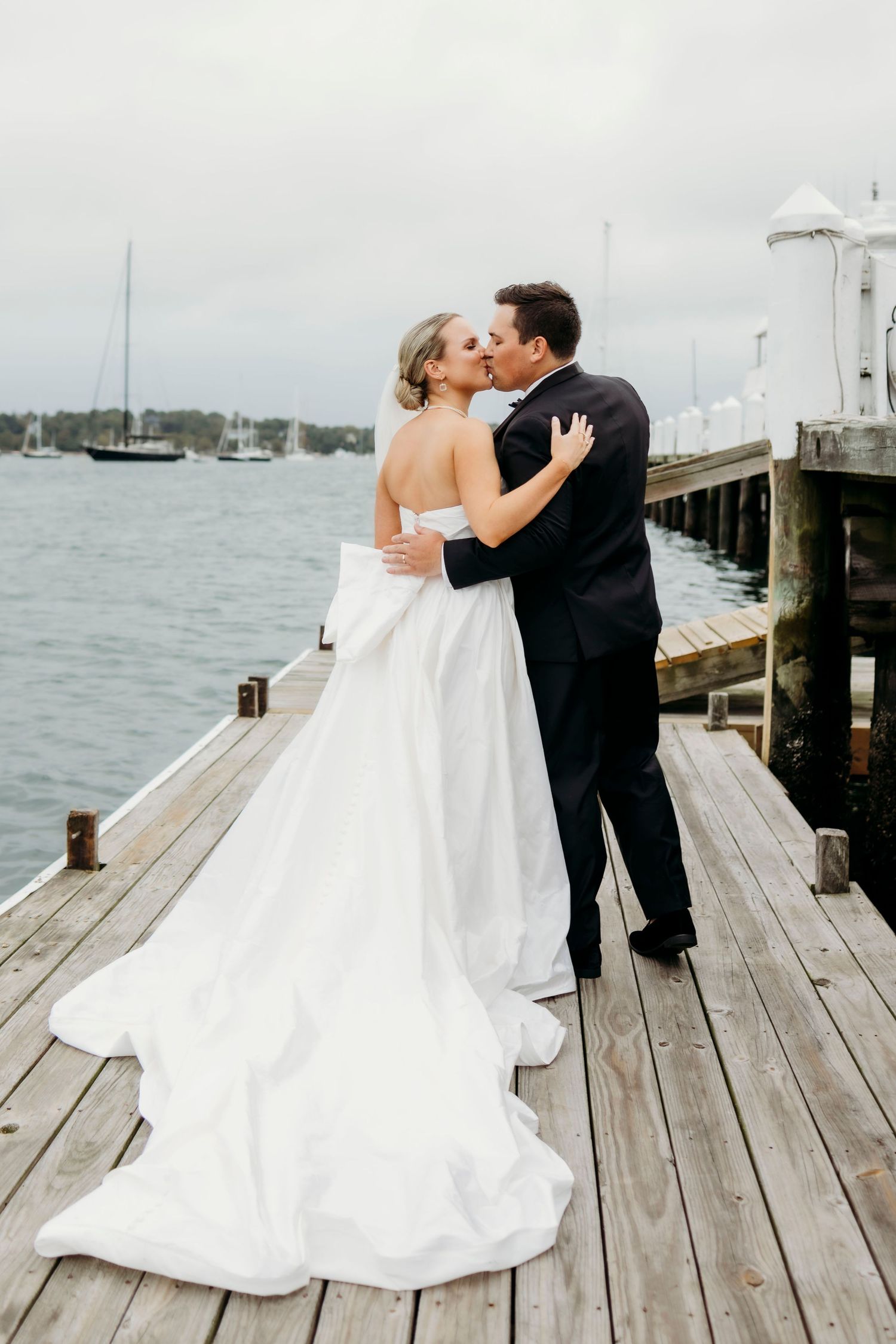 Wedding couple embracing on wooden dock with sailboats in harbor background.
