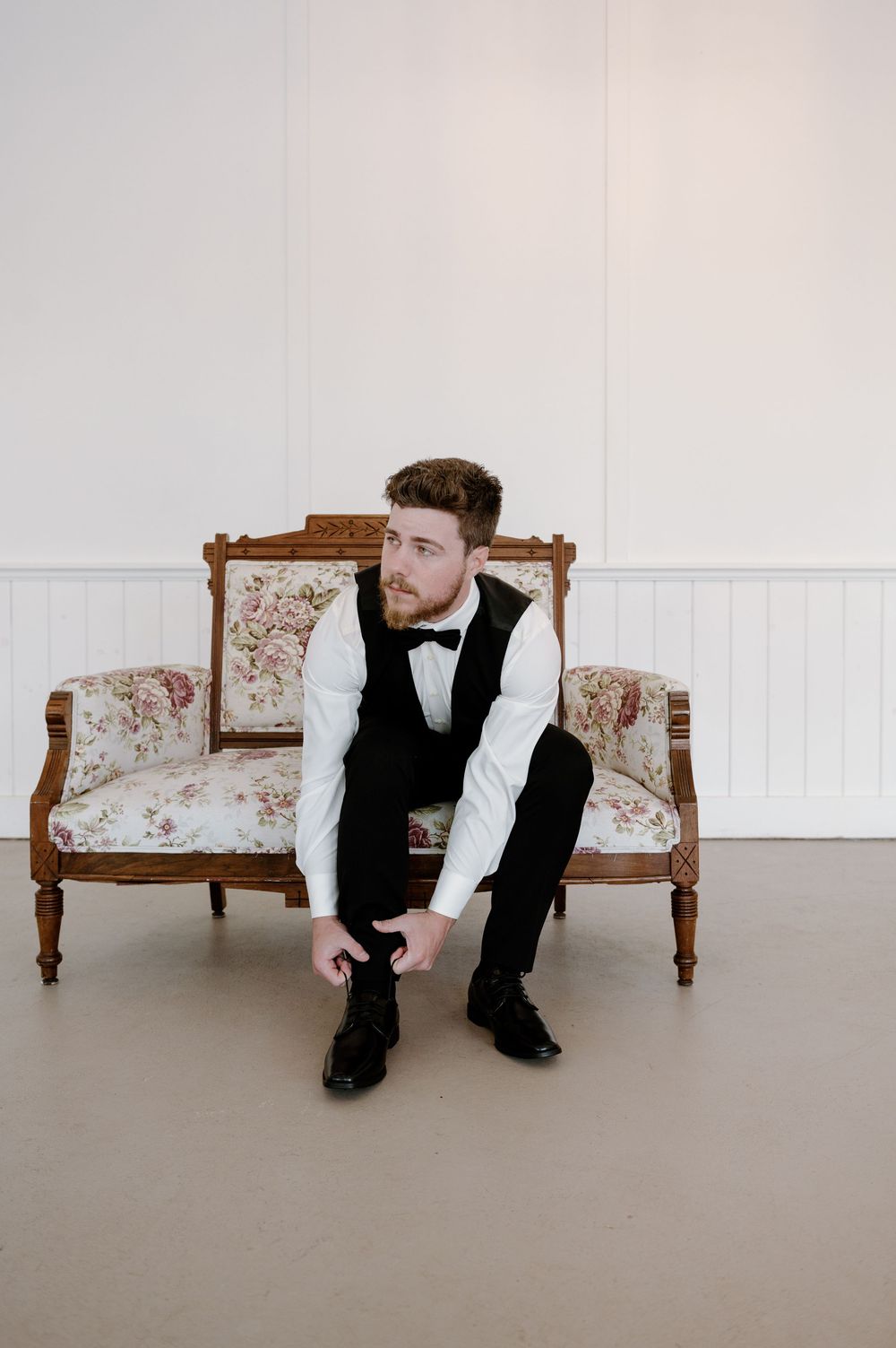 A suited figure sits contemplatively on an antique floral settee in a minimalist setting.