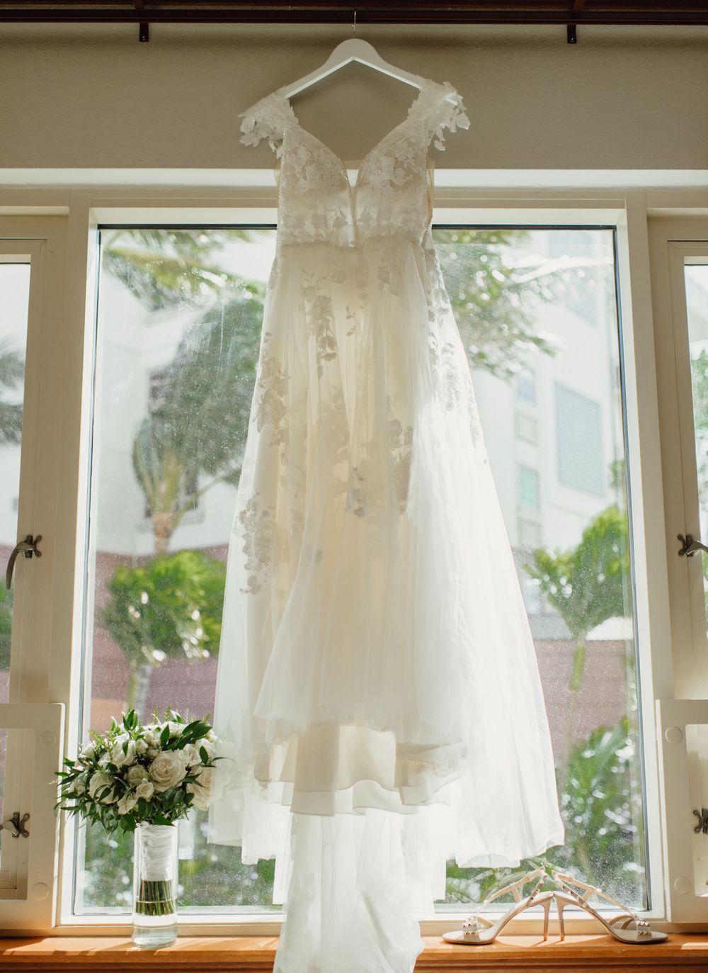 Delicate lace wedding dress hanging in window with bridal bouquet below.