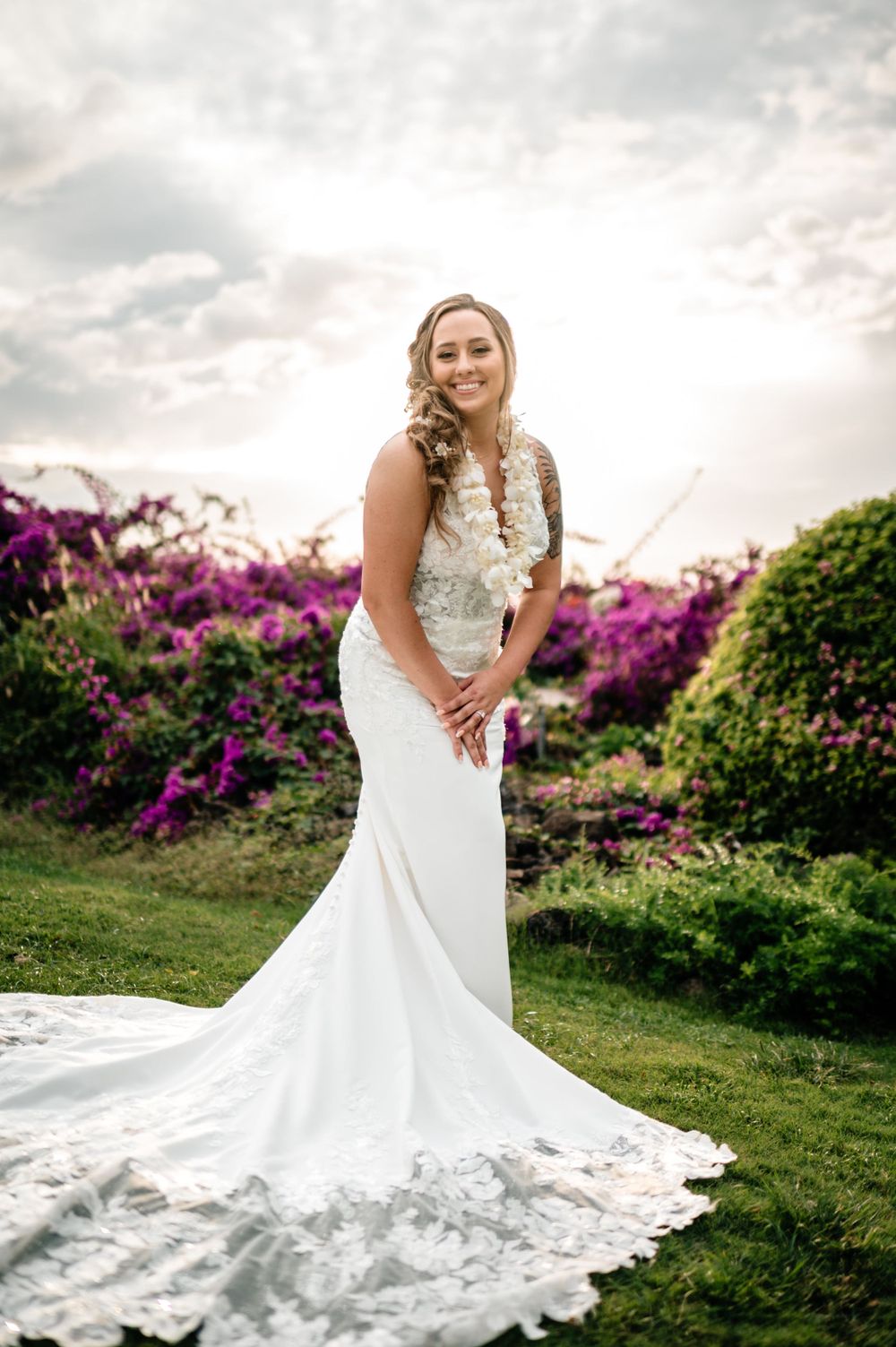 Elegant bride in lace wedding dress poses in garden with purple flowers at sunset.