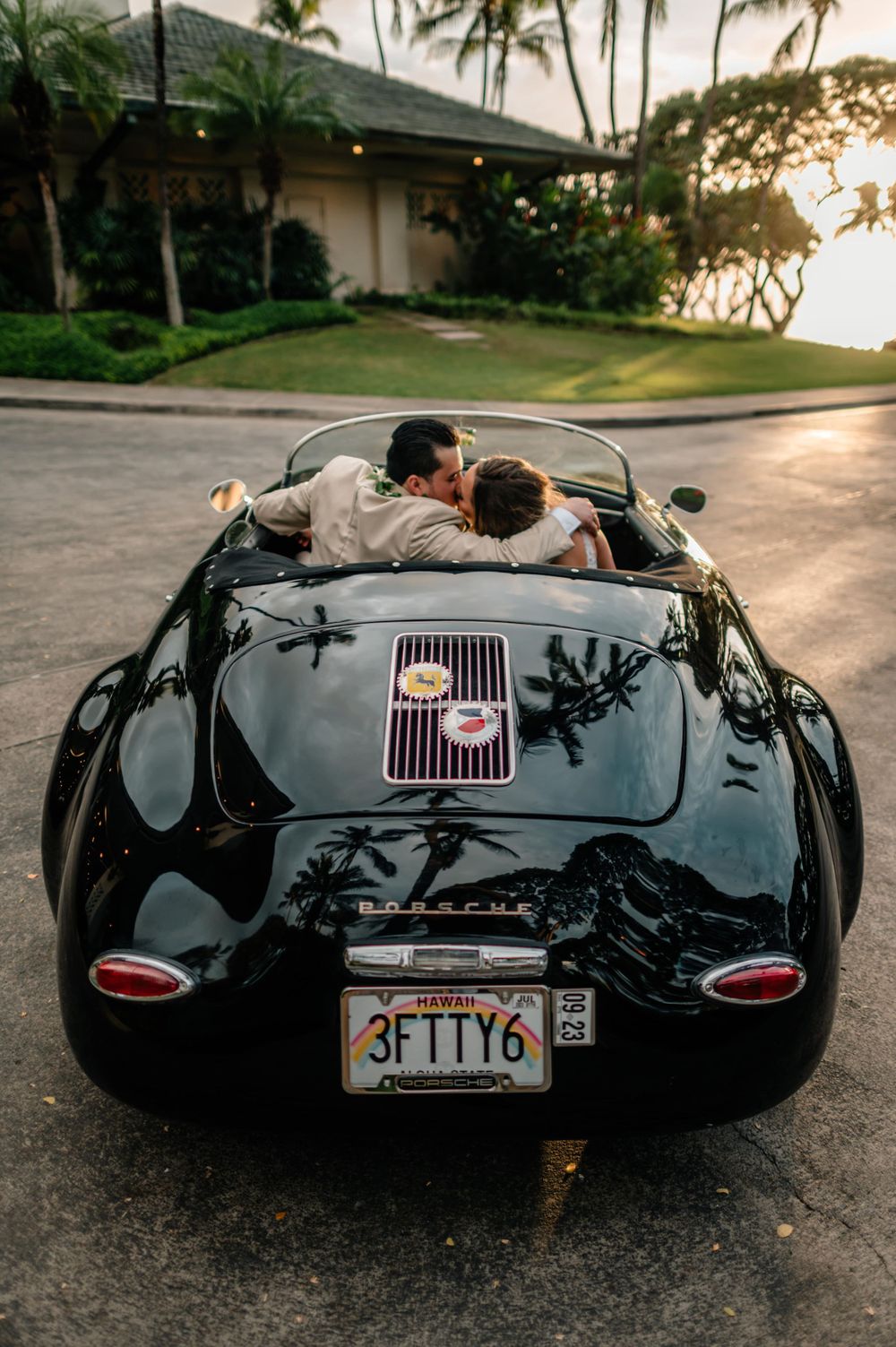 Classic black vintage sports car photographed at sunset with palm trees silhouetted in the background.
