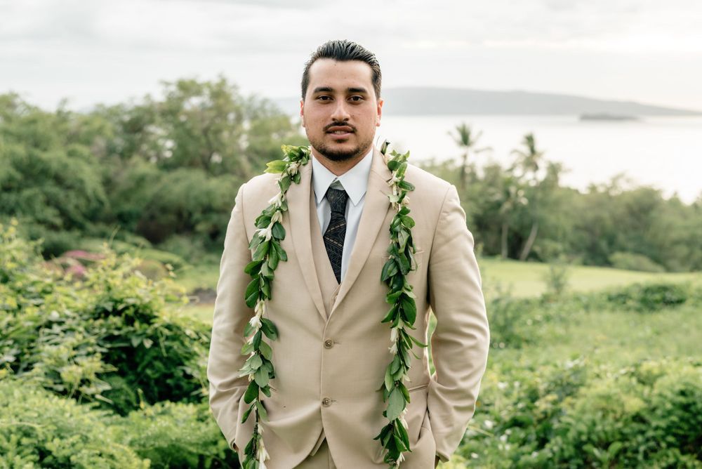 Groom in tan suit with traditional Hawaiian lei stands against tropical landscape.