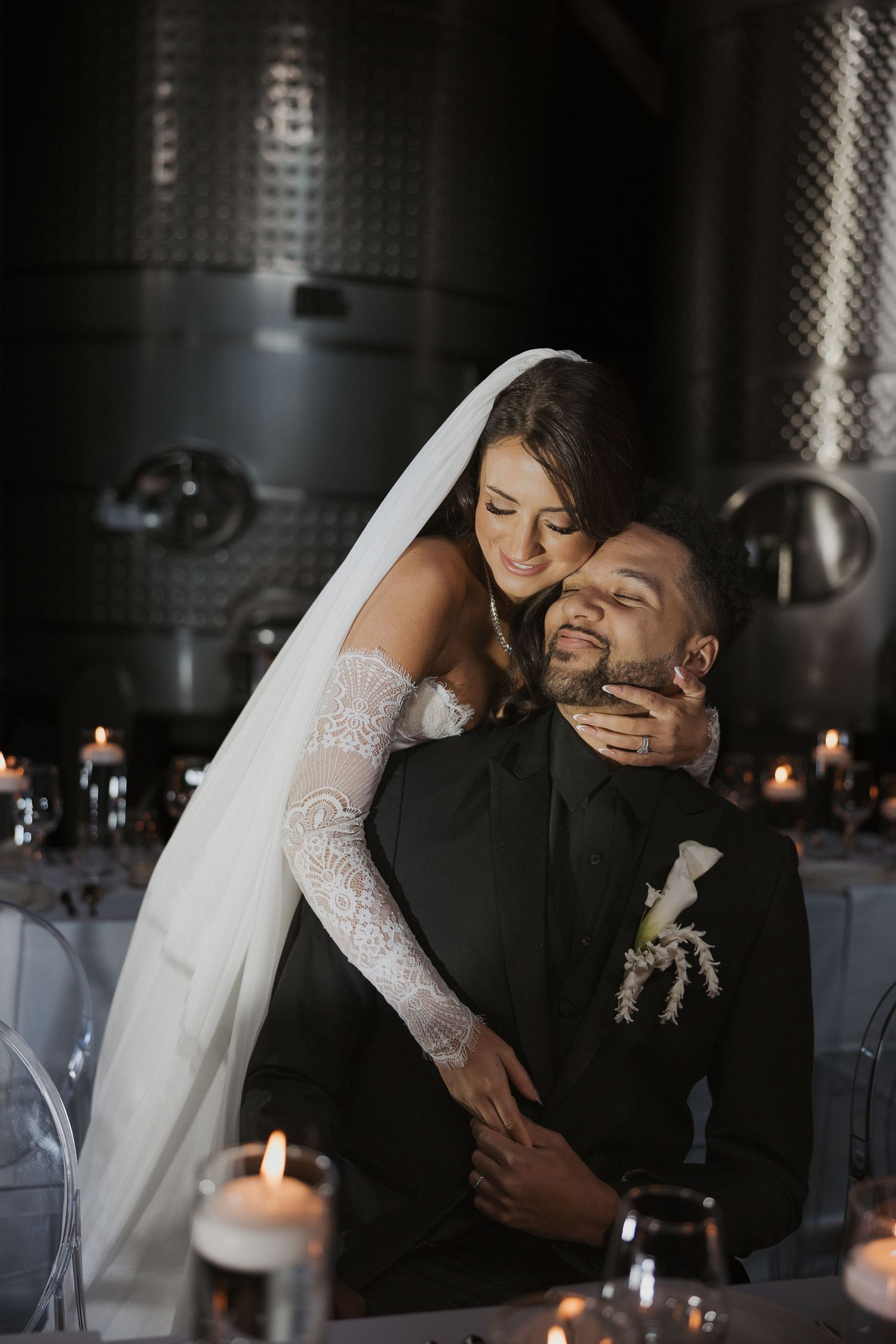 A romantic wedding couple embraces under a flowing white veil against a bright blue sky.