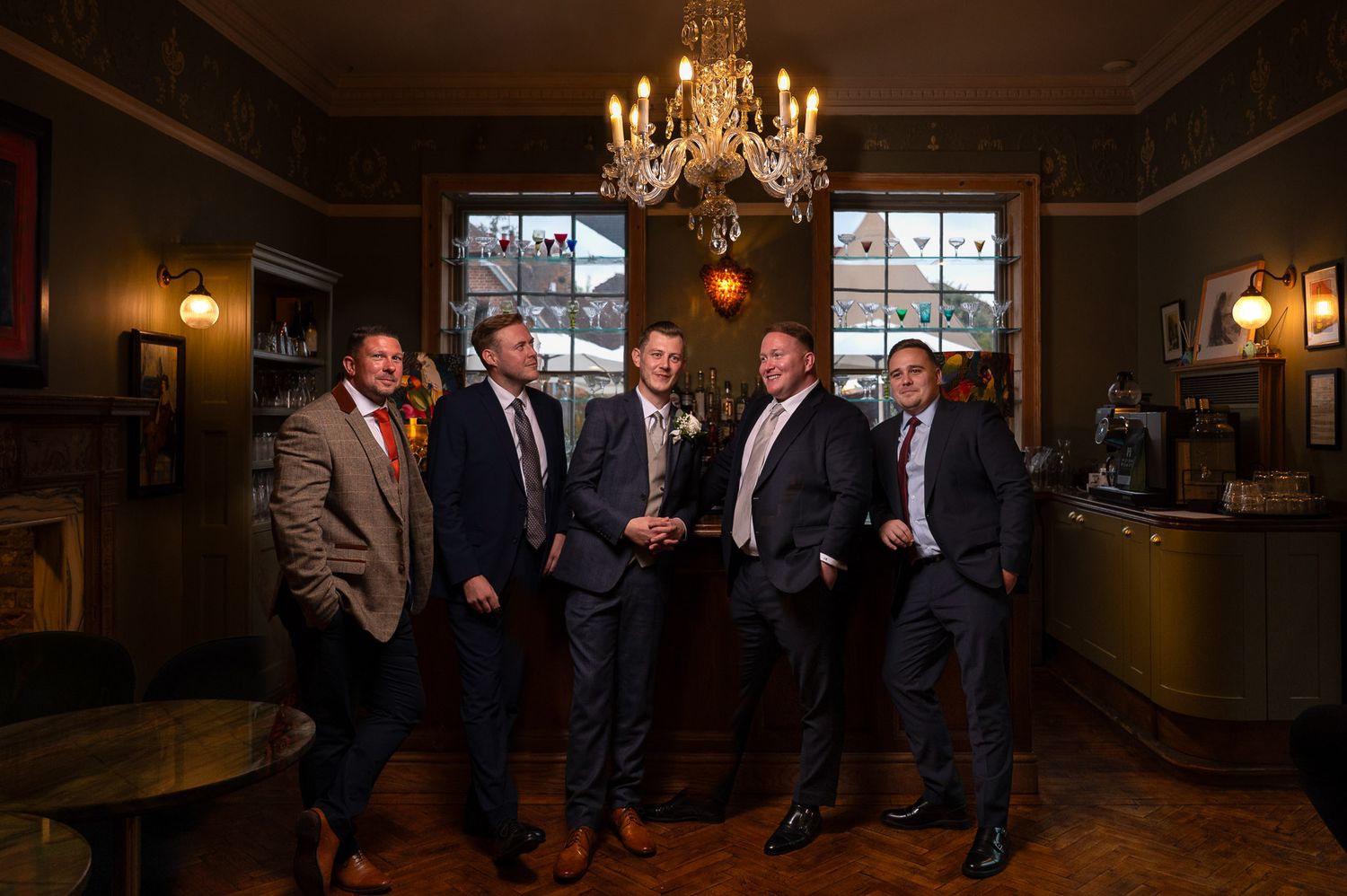 a cinematic portrait of a groom and his groomsmen  in the bar at The Vines Wedding Venue in Rochester. They are lit by warm chandeliers.