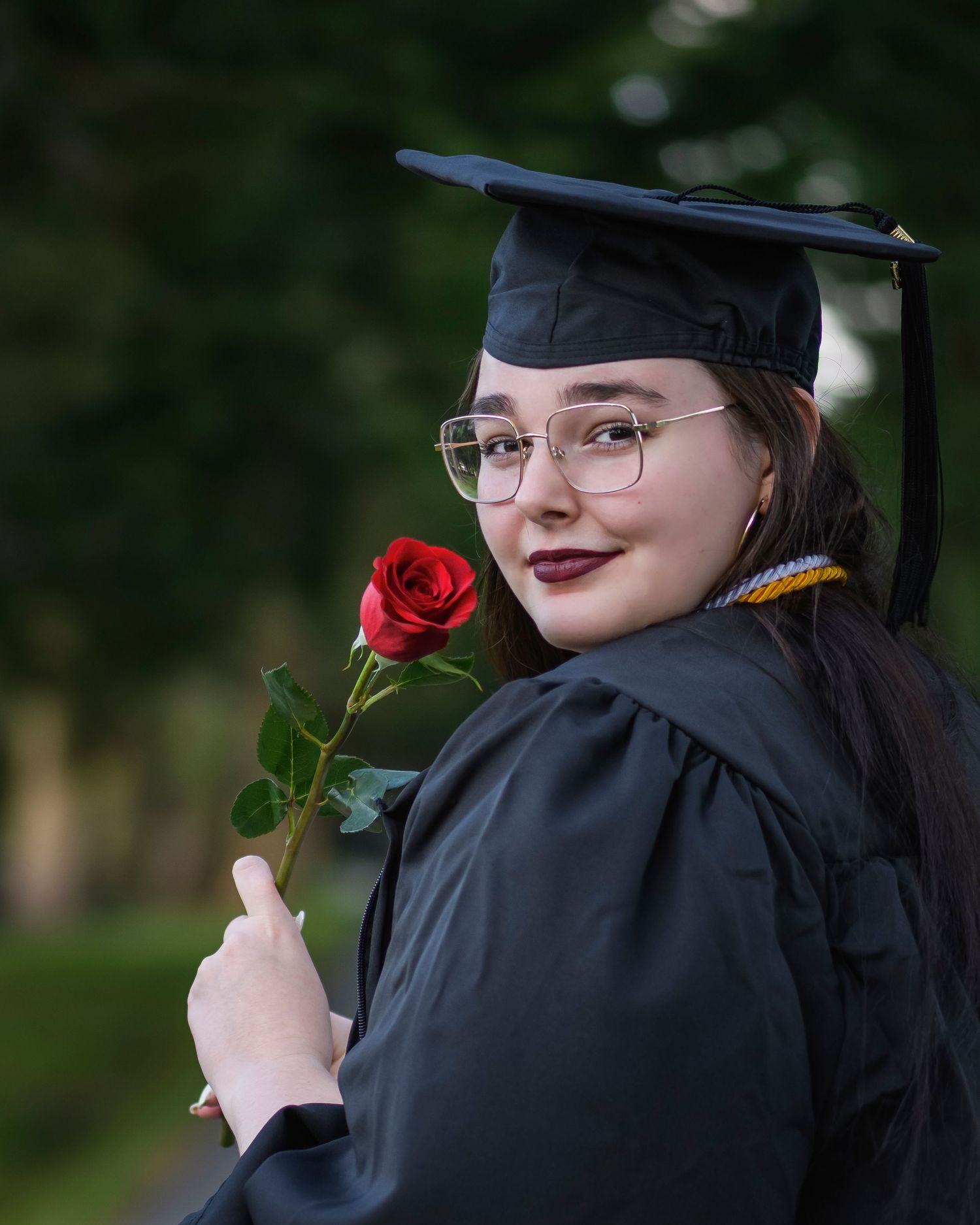 Smiling graduate in black cap and gown holding a red rose, wearing glasses outdoors.