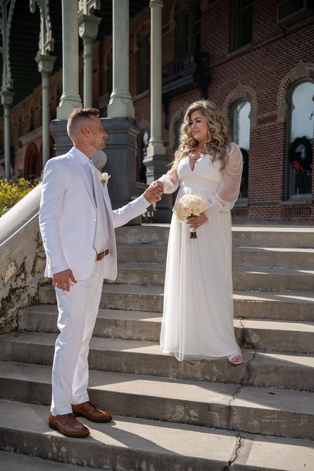 A couple in white attire holds hands on stone steps outside a historic brick building with ornate architecture.