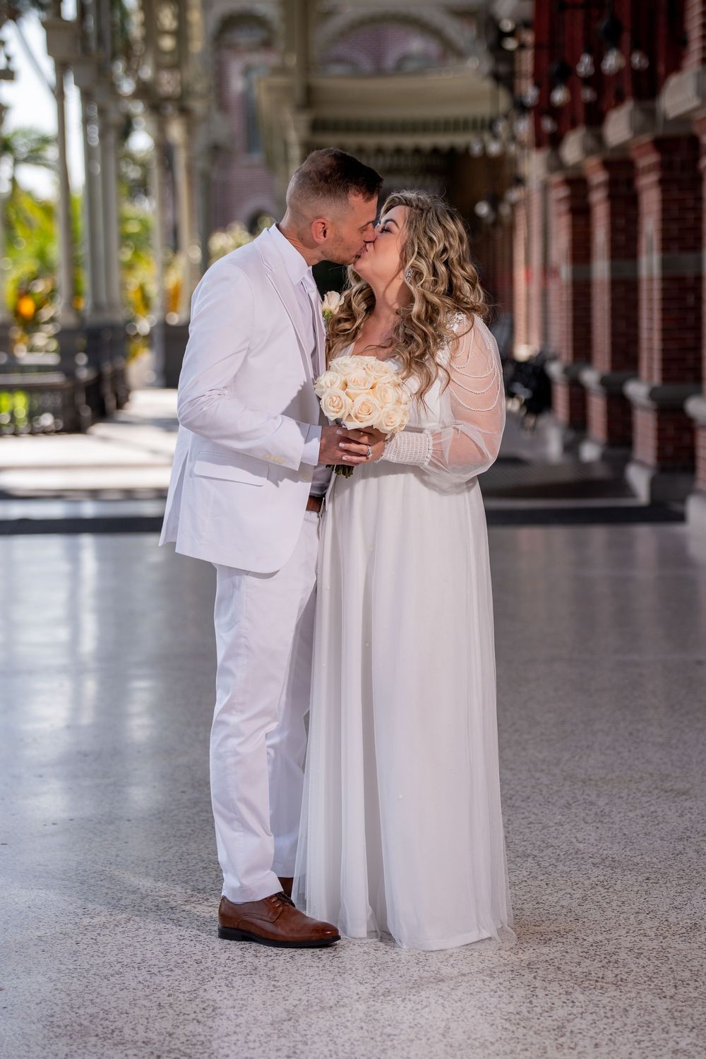 A couple in white attire shares a romantic moment in an elegant columned hallway.