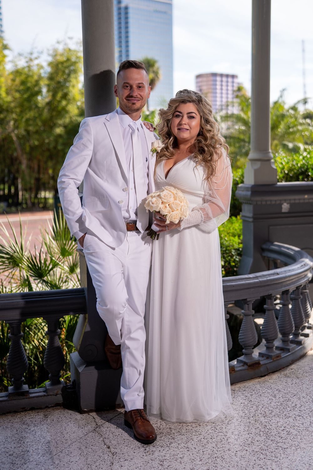 A couple in white attire poses together under a columned gazebo during their wedding celebration.