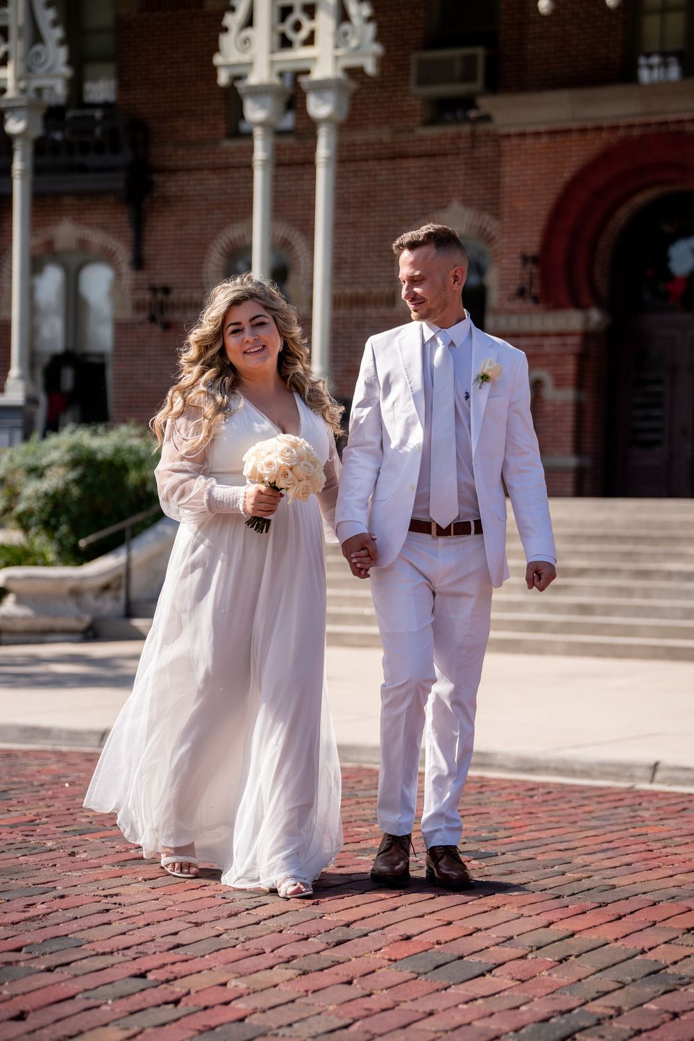 A couple in white wedding attire walks together on red brick pavement outside a historic building.
