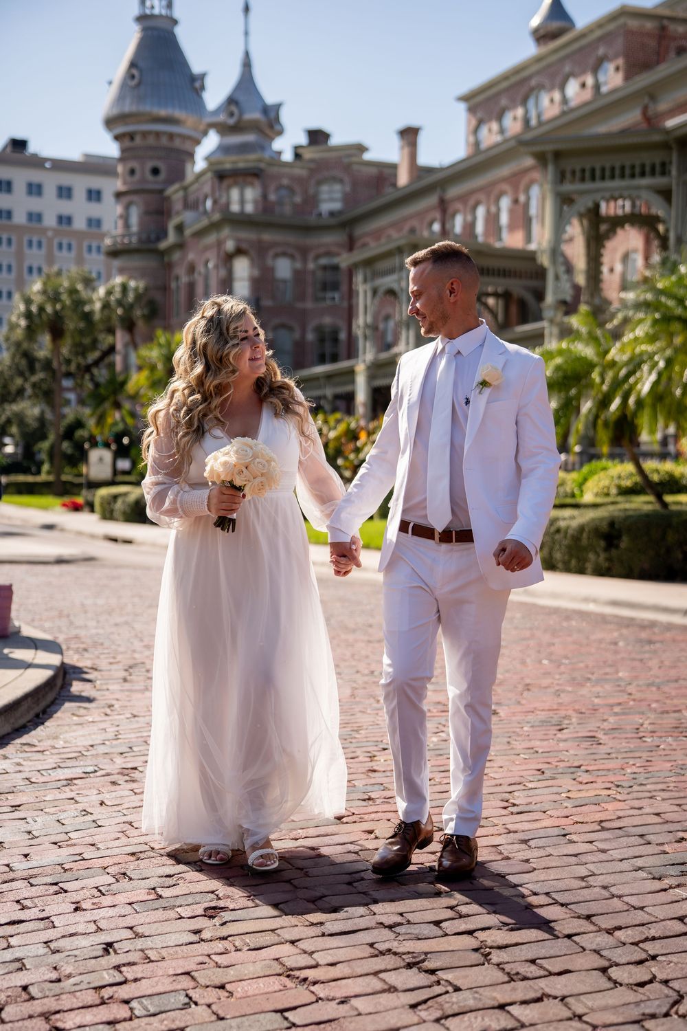 A couple in wedding attire walks along a brick pathway with ornate buildings in the background.