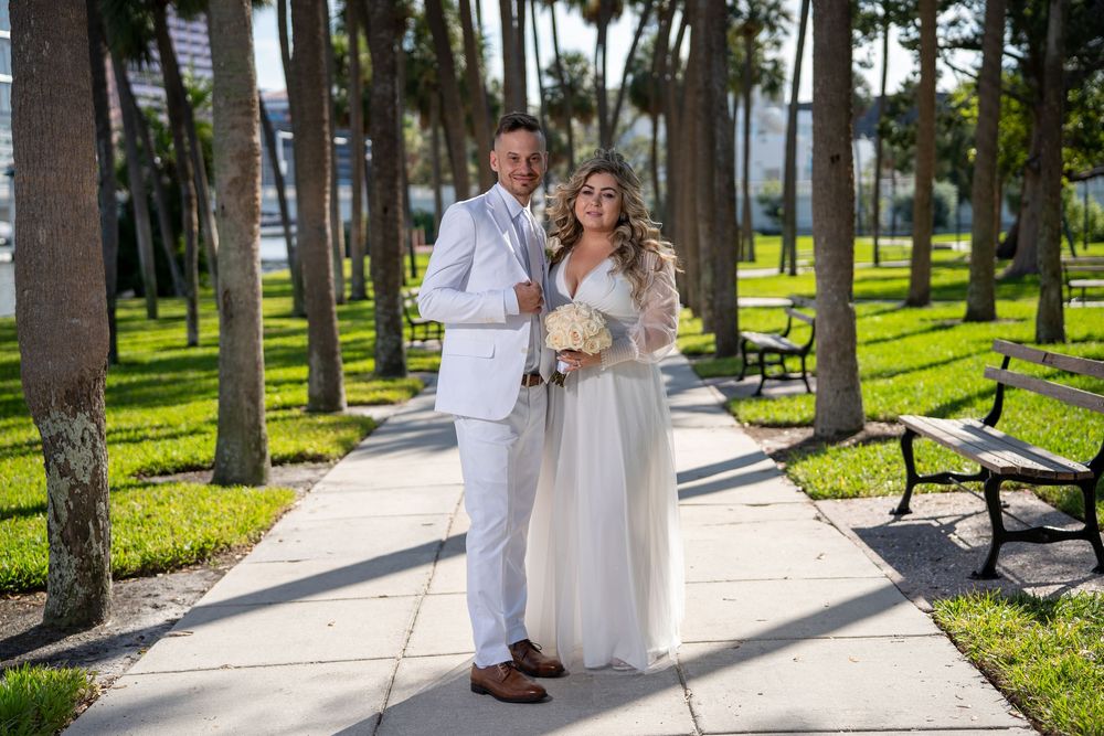 A couple in white attire embraces while walking along a sunlit path lined with tall pine trees in a park.