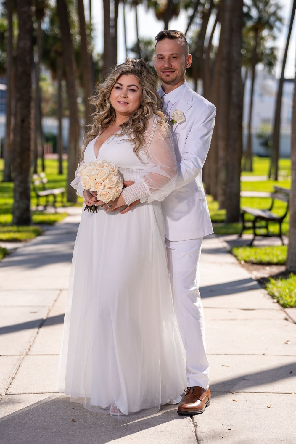A couple in white wedding attire poses together on a palm tree-lined path during a sunny outdoor photoshoot.