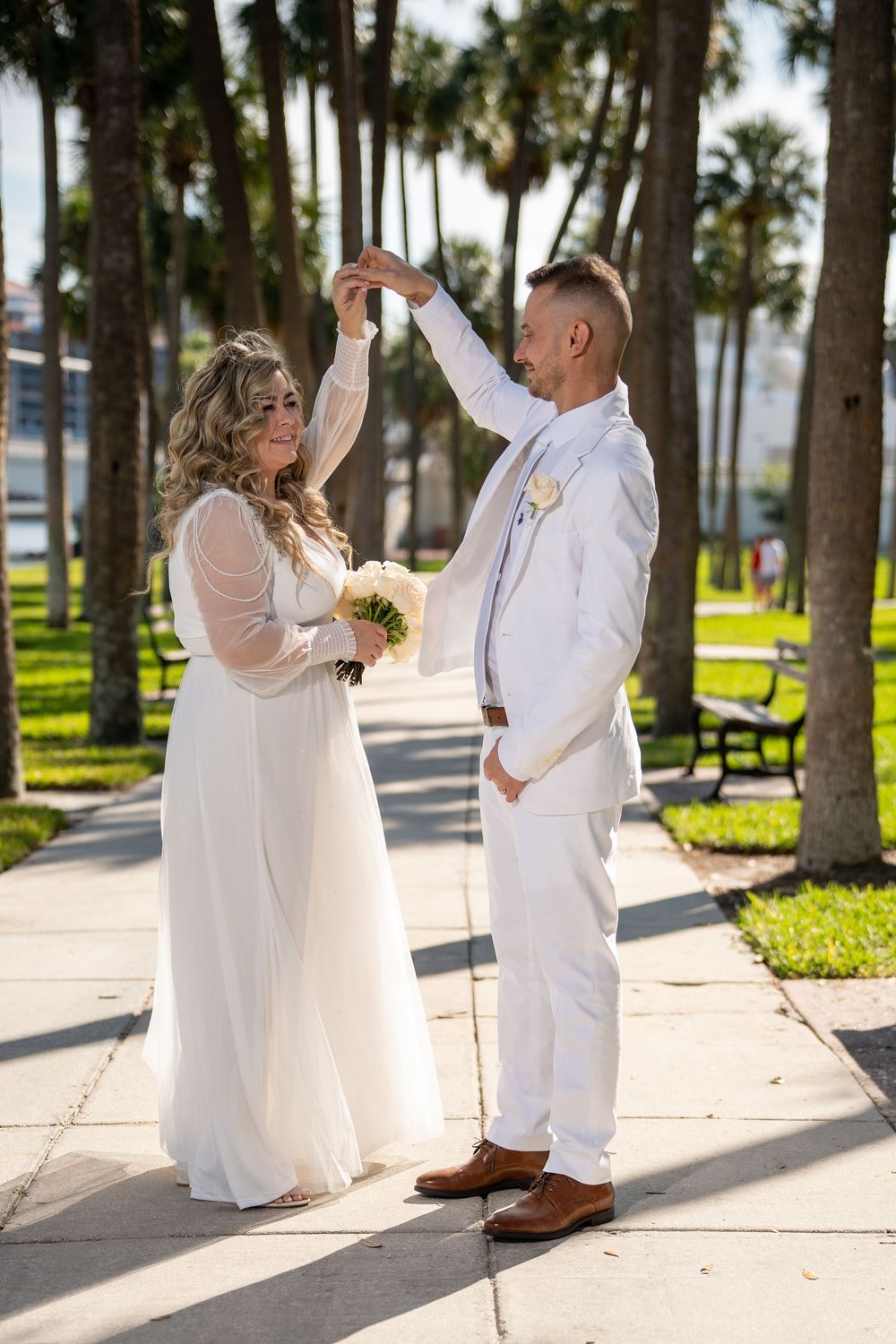 A couple in white attire dances together on a palm tree-lined path during a romantic beach wedding photoshoot.