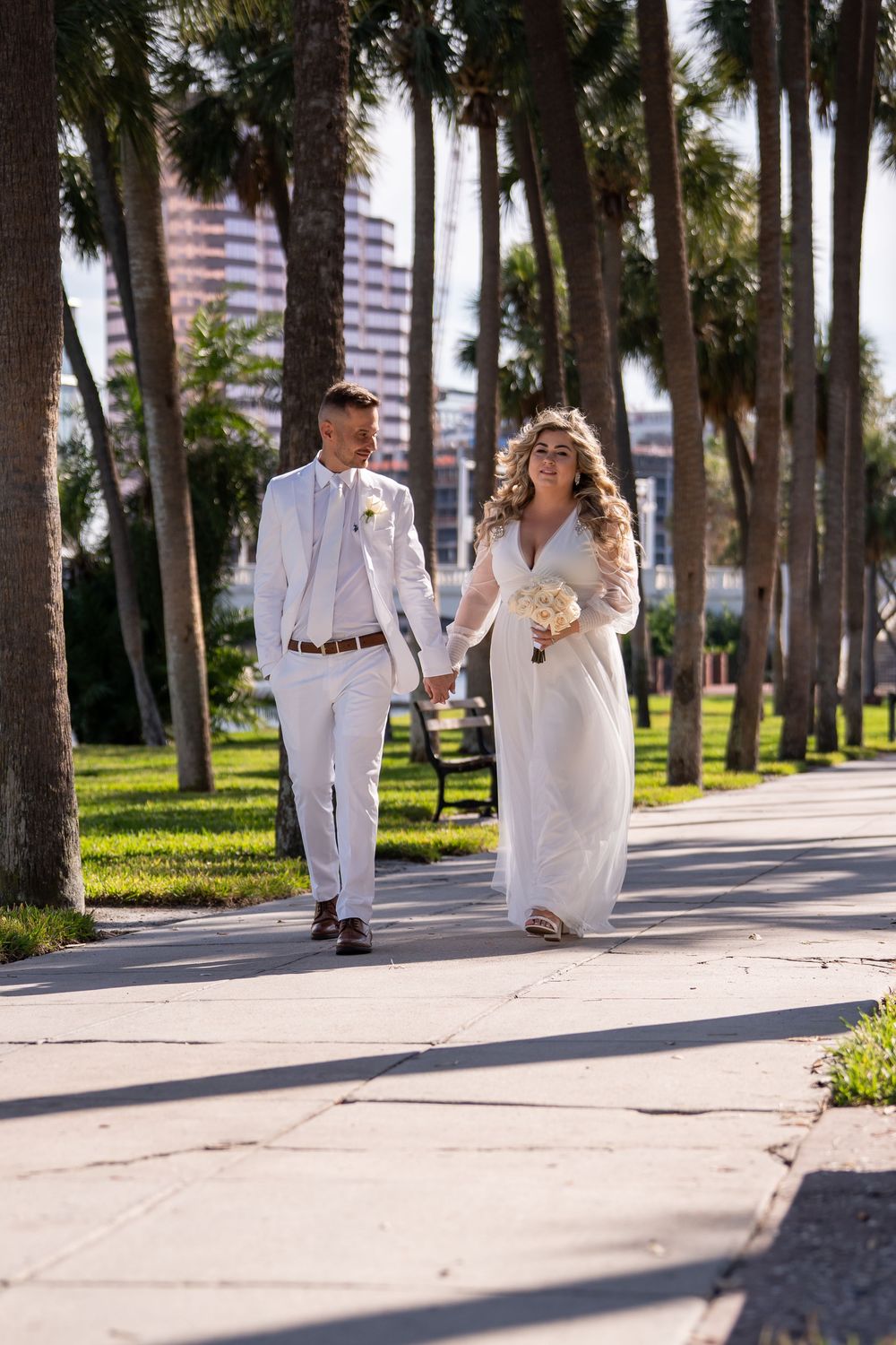 A couple in white outfits walks along a palm tree-lined pathway in a tropical setting.