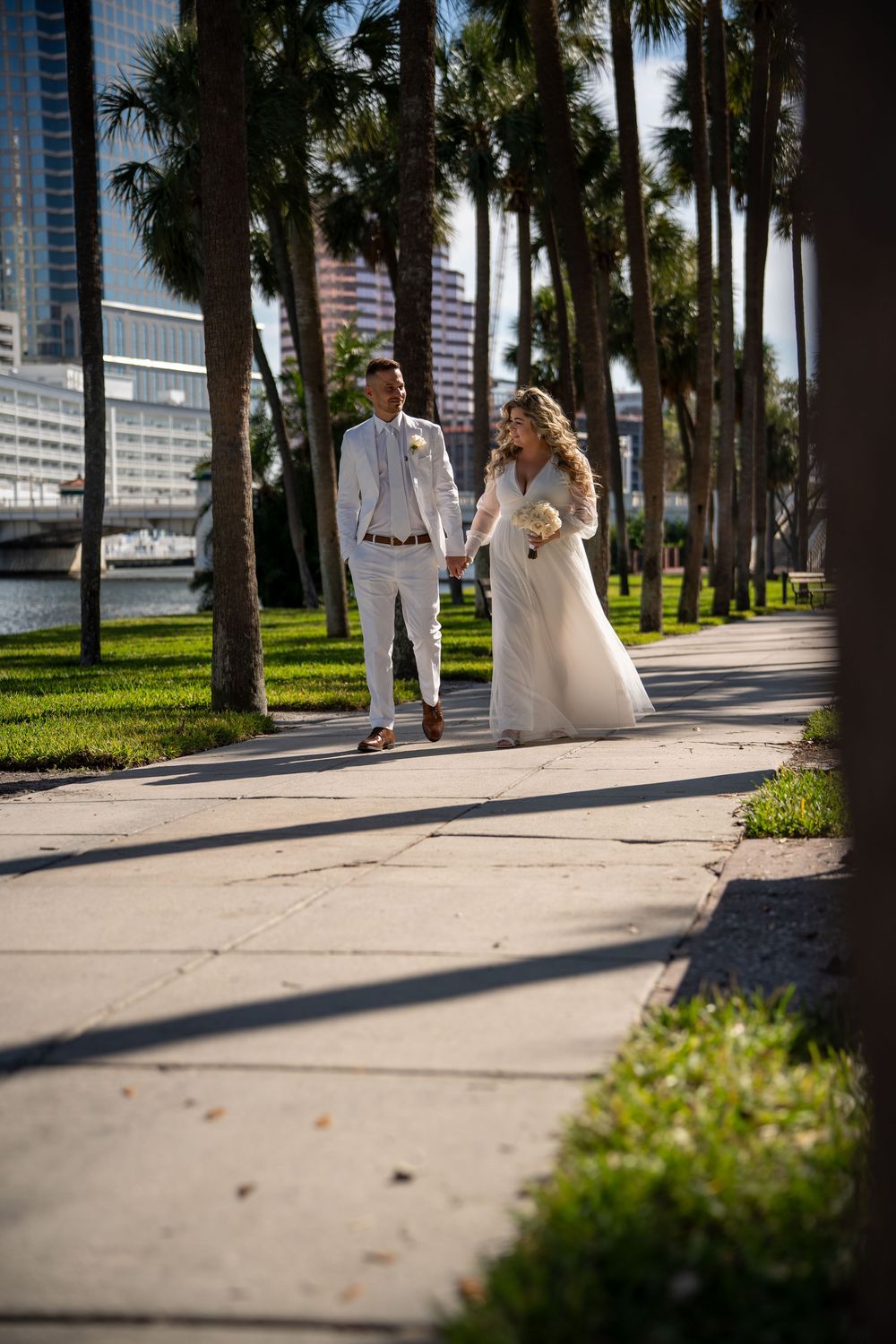 Wedding couple walks along palm-lined pathway in Miami with downtown skyline and waterfront in background.