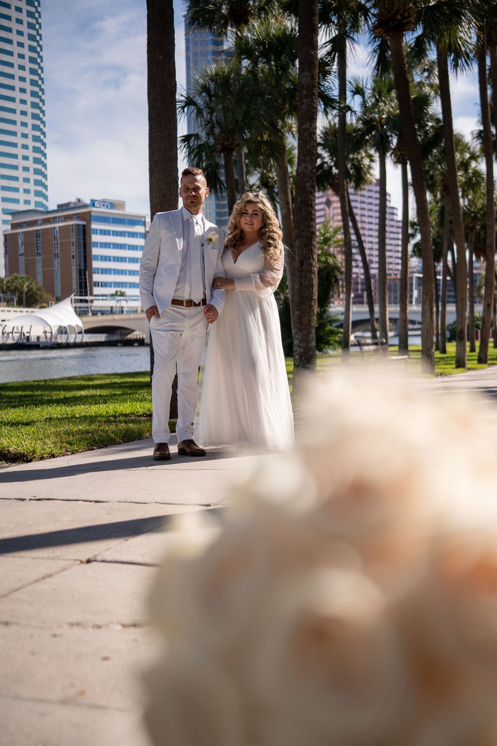 A couple in white wedding attire walks along a palm-lined path with modern city buildings in the background.