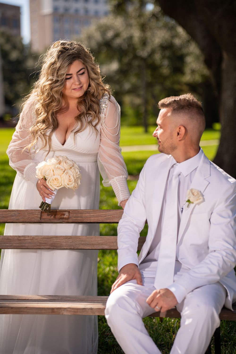 A romantic wedding scene outdoors with the couple in white attire sitting on wooden steps holding a bouquet of roses.