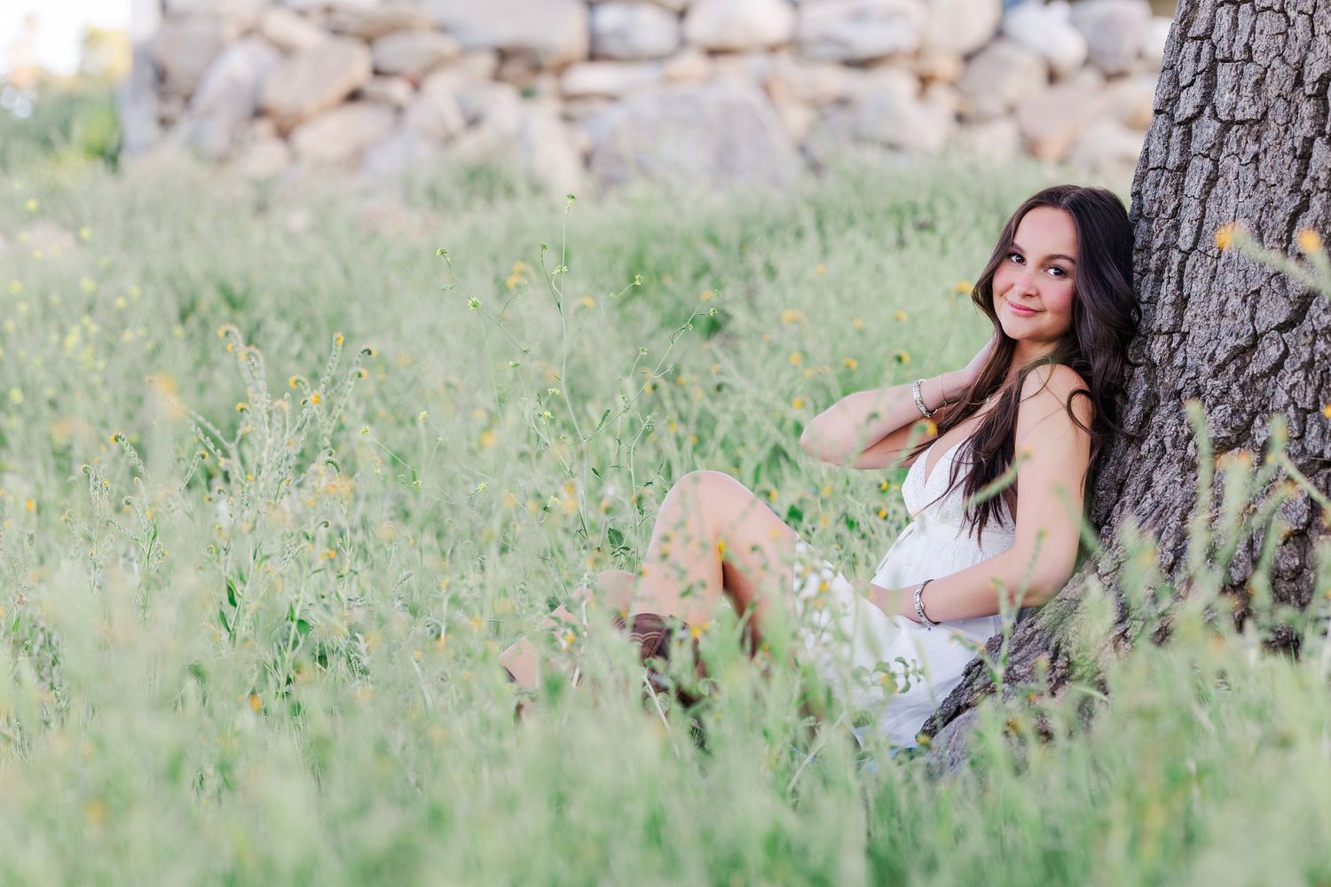 A person in a white dress poses joyfully among tall green grass and wildflowers in a sunny outdoor setting.
