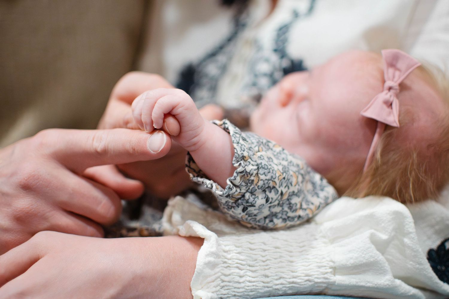 Newborn baby in floral outfit with pink bow gently holding parent's finger.
