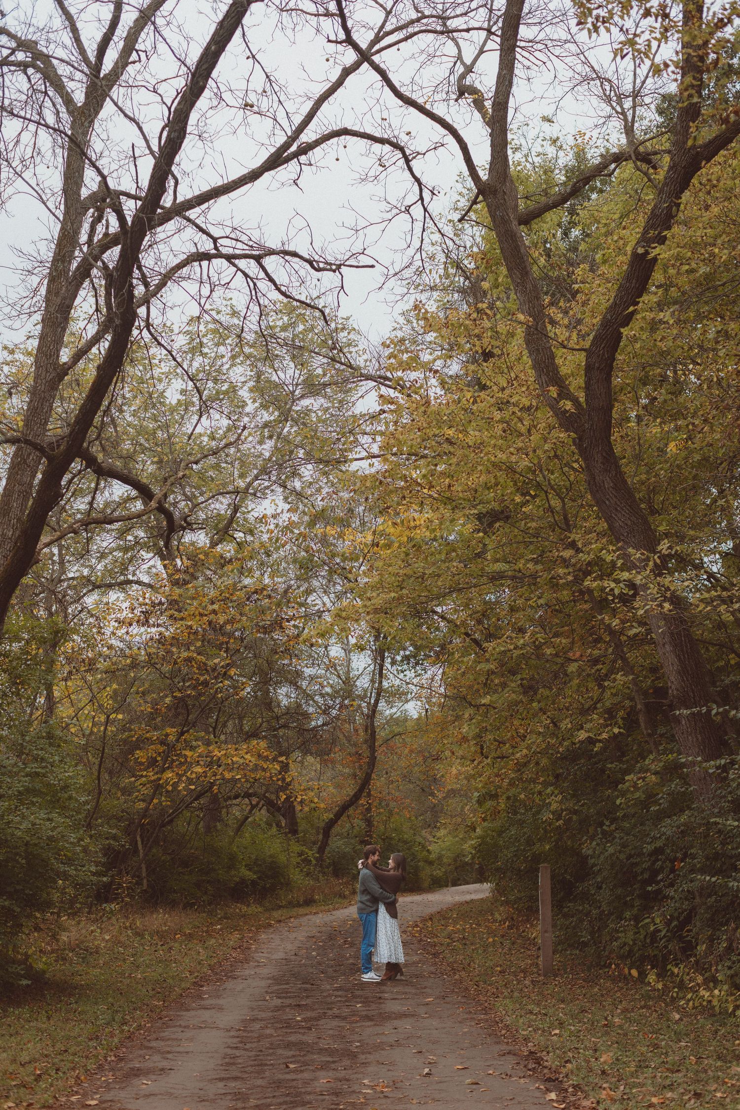 A pulled-back shot of a man and woman standing close on a tree-lined path during an engagement photo session in Kansas City, Missouri.