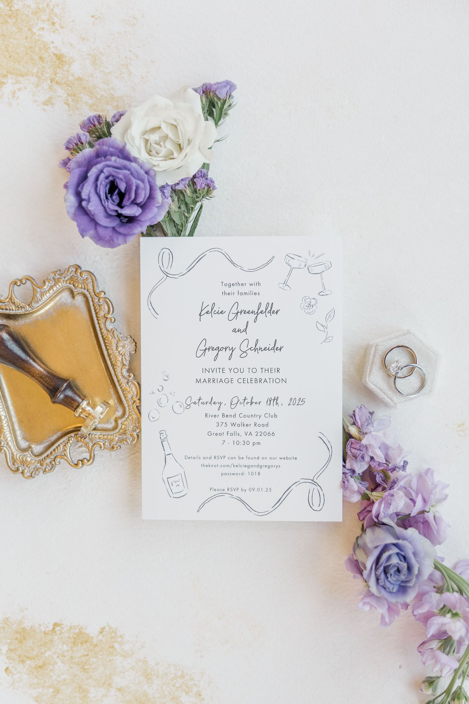 Wedding invitation suite with purple flowers and gold accents laid out on white surface with wedding rings.