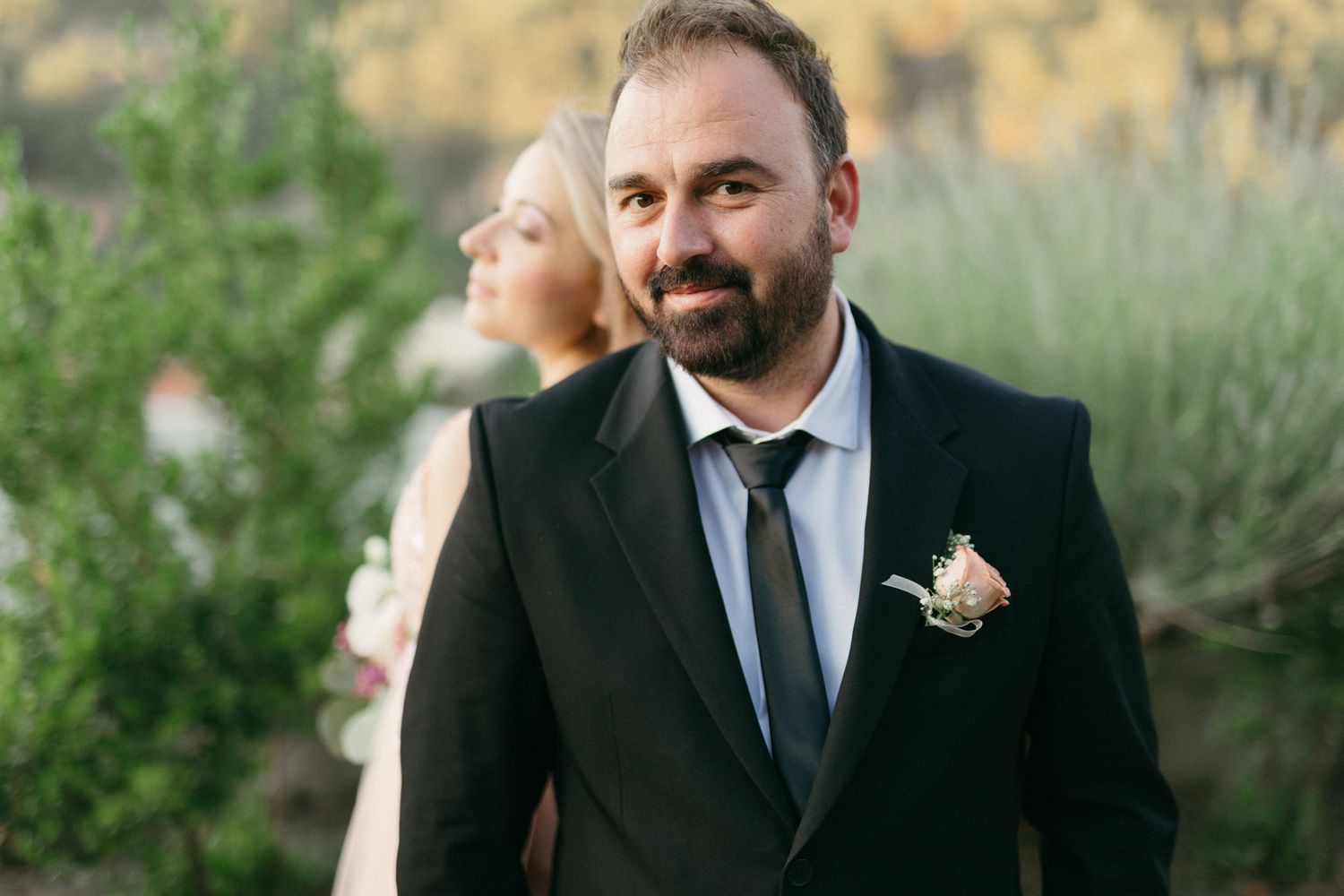 A wedding party stands together outdoors in formal attire with greenery and natural lighting in the background.