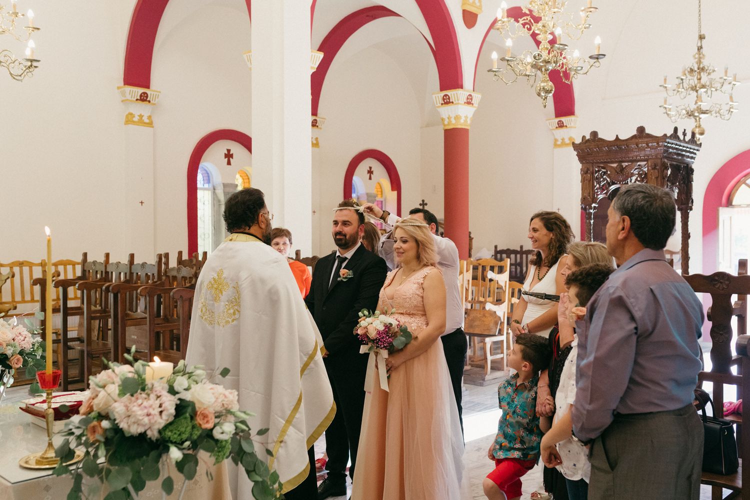Wedding party gathers in elegant church venue with white floral arrangements and red accents throughout the space.