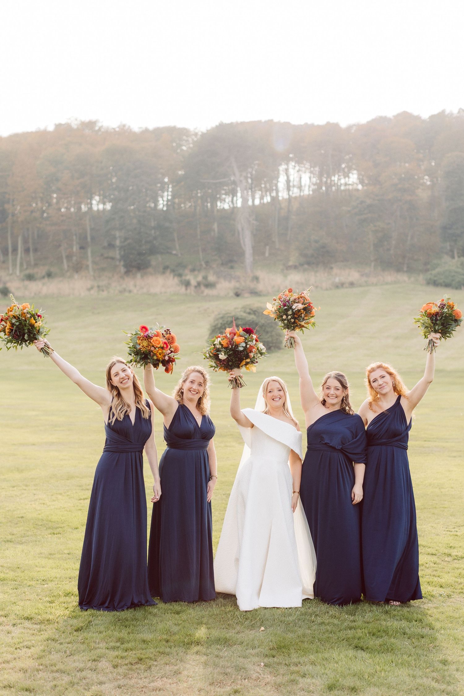 Bridesmaids in navy dresses holding autumn bouquets celebrating in sunlit field.