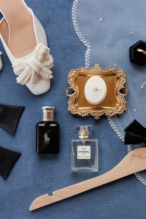 Wedding accessories including perfume bottles, pearl necklaces, white shoes, and black bow ties arranged on blue fabric.
