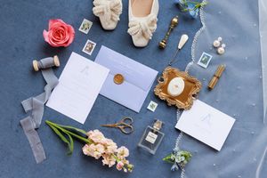 A flat lay composition of wedding details including pink roses, ribbons, invitations, and vintage styling elements on blue surface.