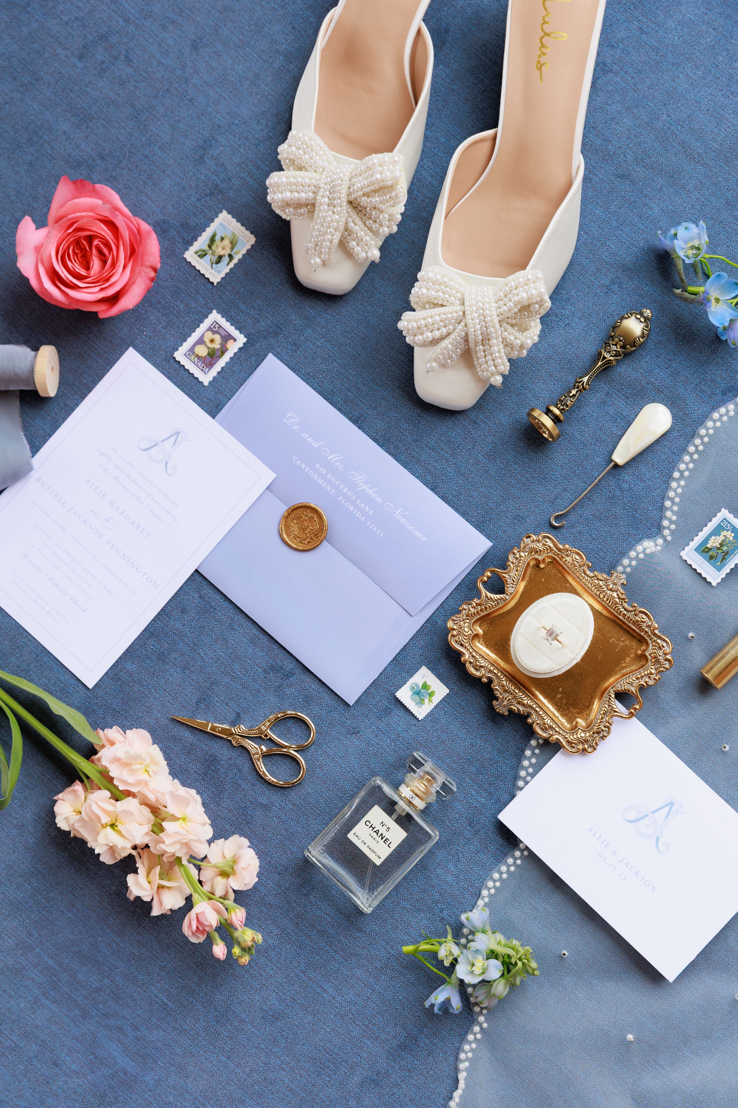 Flat lay of wedding accessories including ivory shoes, flowers, candles, and stationery on blue background.