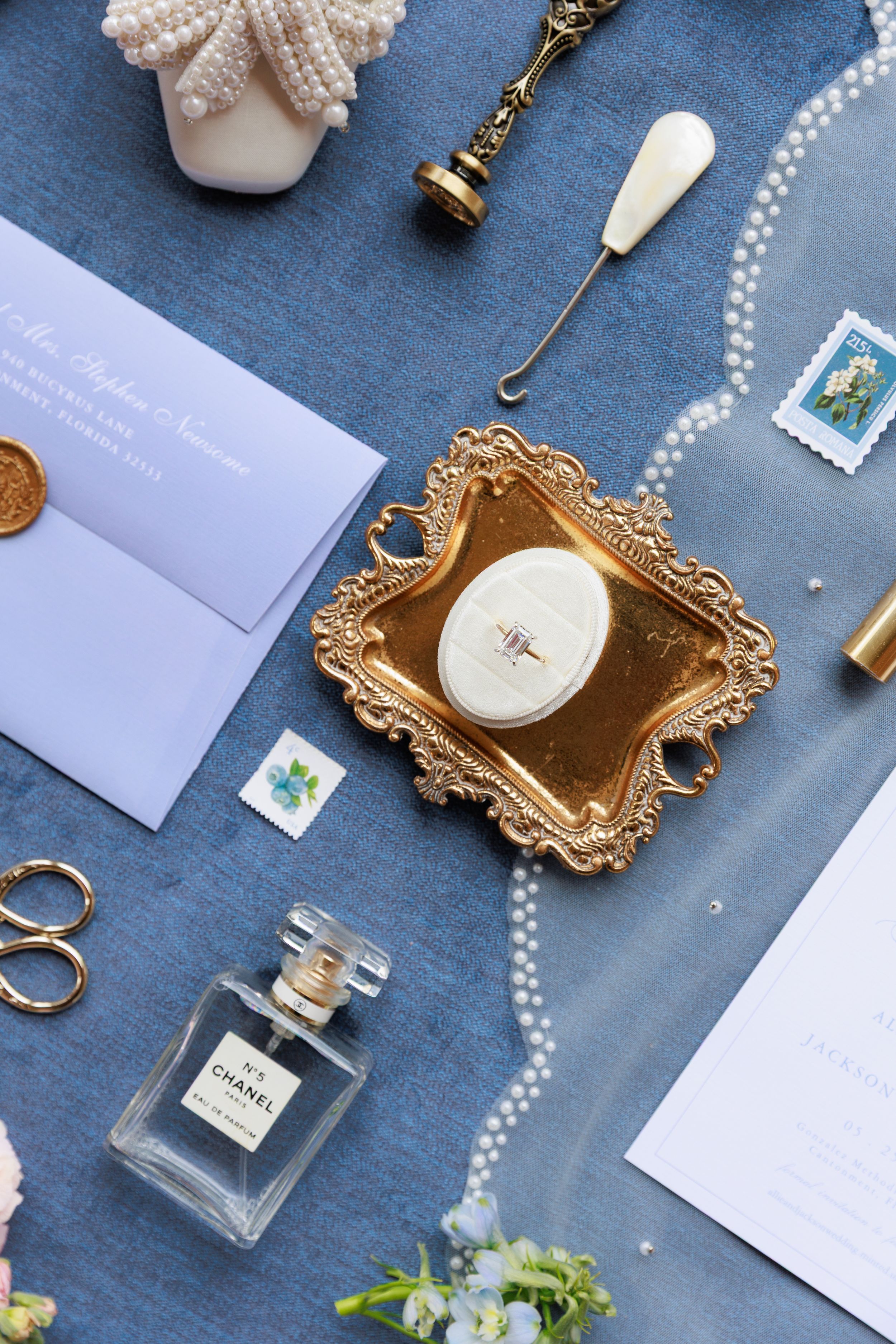 Wedding accessories including perfume bottles, pearl necklaces, seashells and invitations arranged on blue fabric.